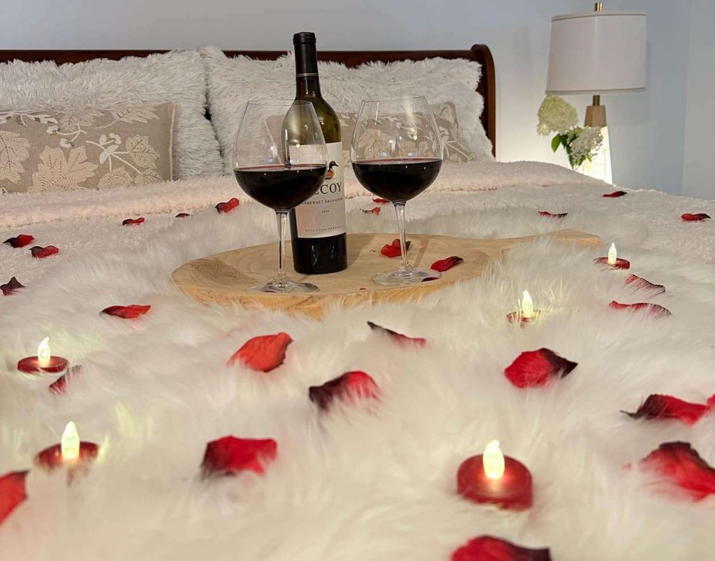 Two wine glasses, bottle of wine, rose petals, and tea lights on a bed